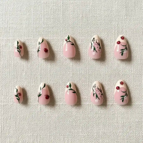 Handmade press-on nails in soft pink and nude tones with hand-painted ladybug, red berry, green leaf, and white flower nail art, almond shape, 10 pieces, flat lay on linen background